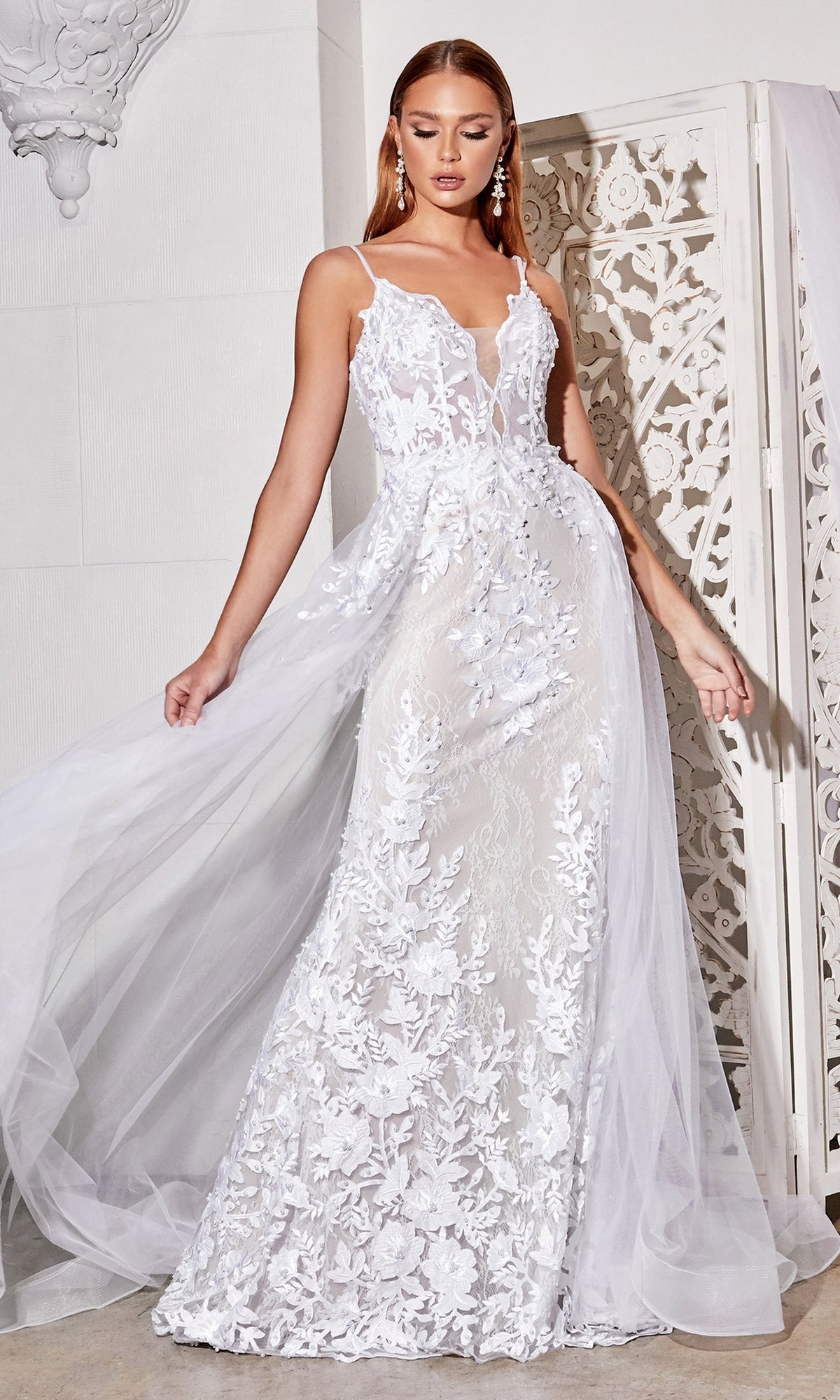 A woman stands indoors near a decorative white screen wearing the Sheer-Corset Long White Lace Wedding Dress CD931W, featuring sleeveless design, floral embroidery, and a flowing sheer overskirt.